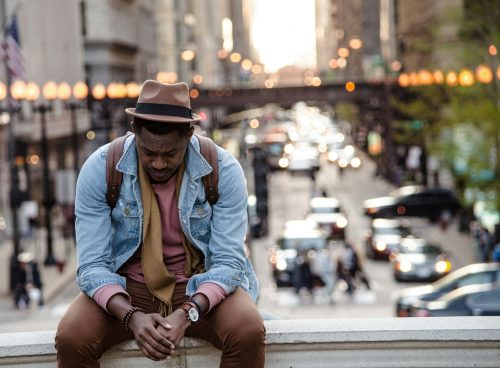 A man in a leather jacket looking down while sitting on a ledge in a city
