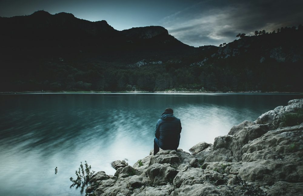 a man sits on a rock staring across a lake at dusk