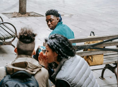 Back view of faceless classmates spreading gossip about African American man on wooden bench