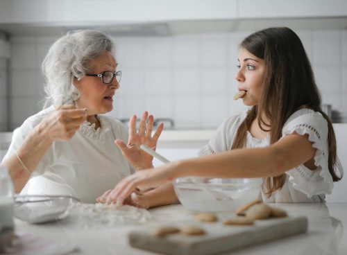 Calm senior woman and teenage girl in casual clothes looking at each other and talking while eating cookies and cooking pastry in contemporary kitchen at home