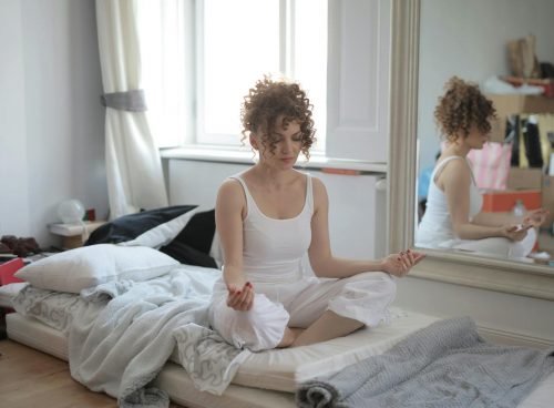 Tranquil female in sleepwear sitting on bed in messy room and practicing yoga with closed eyes while maintaining mental health