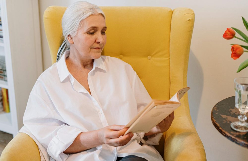 An Elderly Woman Sitting On A Couch Reading A Book