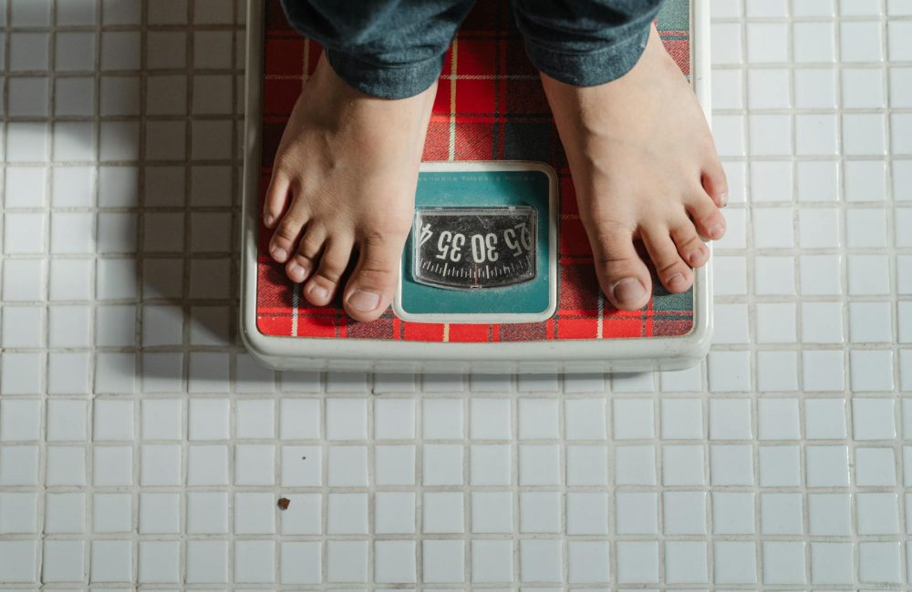 From above crop anonymous barefoot child in jeans standing on weigh scales on tiled floor of bathroom