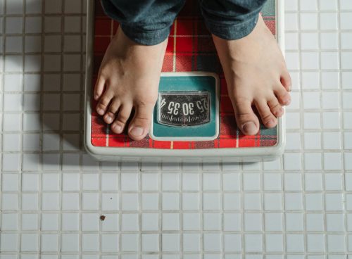 From above crop anonymous barefoot child in jeans standing on weigh scales on tiled floor of bathroom