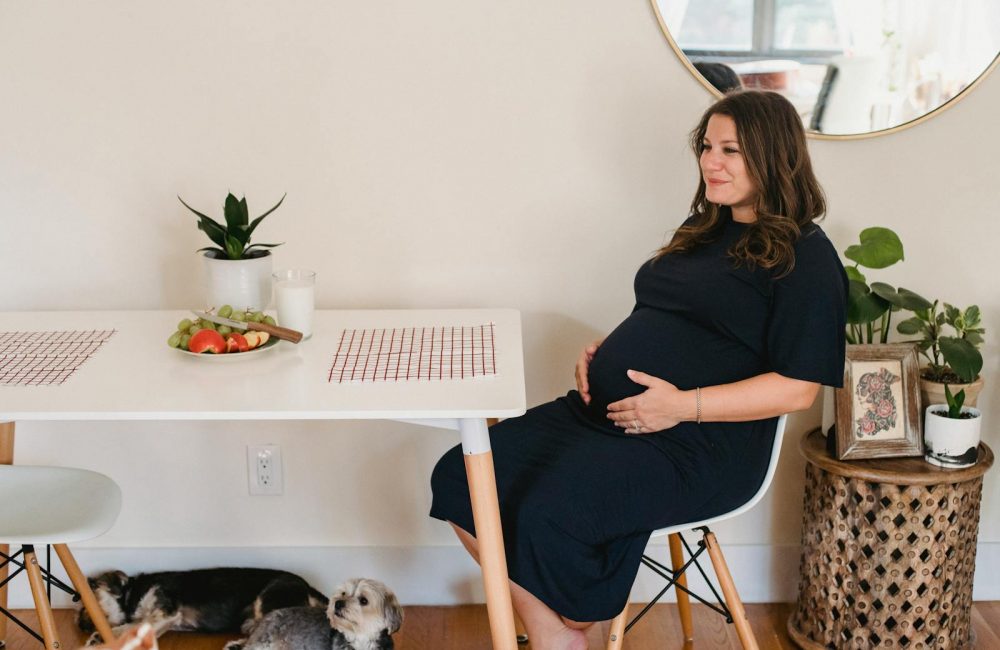 Side view of positive pregnant female in casual clothes touching belly and sitting at table while chilling at home with dogs in daytime