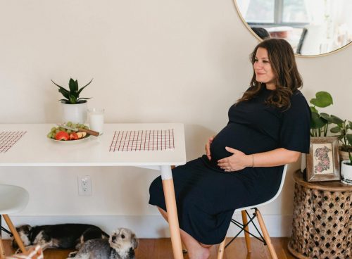 Side view of positive pregnant female in casual clothes touching belly and sitting at table while chilling at home with dogs in daytime