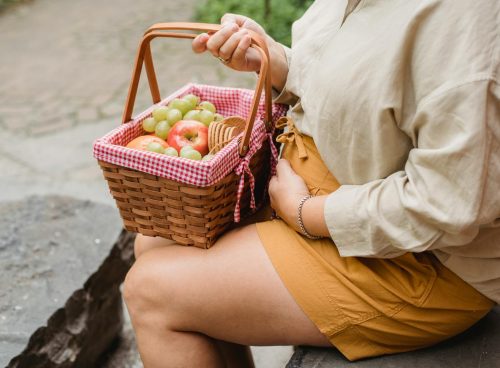 Crop anonymous female in expectancy wearing casual clothes with basket full of ripe fruits and biscuits prepared for picnic