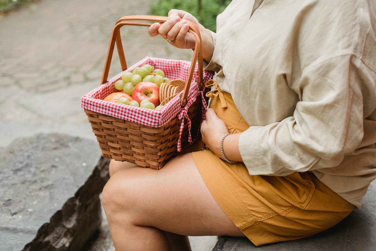 Crop anonymous female in expectancy wearing casual clothes with basket full of ripe fruits and biscuits prepared for picnic