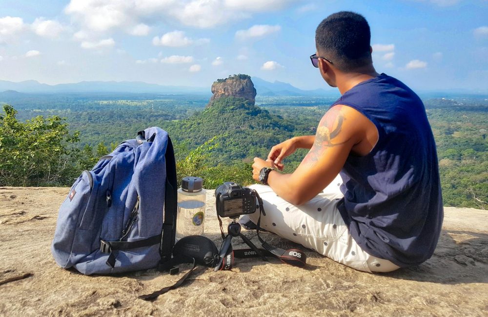 Man Sitting on Top of Gray Cliff Mountain Beside Backpack, Water Bottle, and Camera