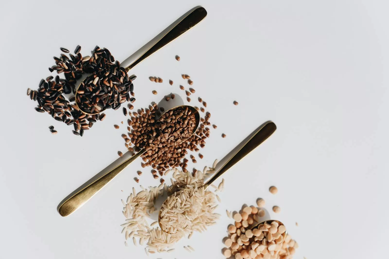 Top view of various dry grains on spoons against a white background, showcasing a variety of textures and colors.